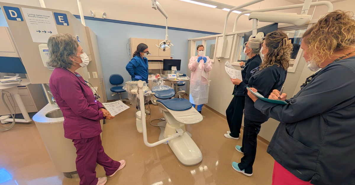 Photo of dental hygienists aruond a dental chair room with a woman leading the course.